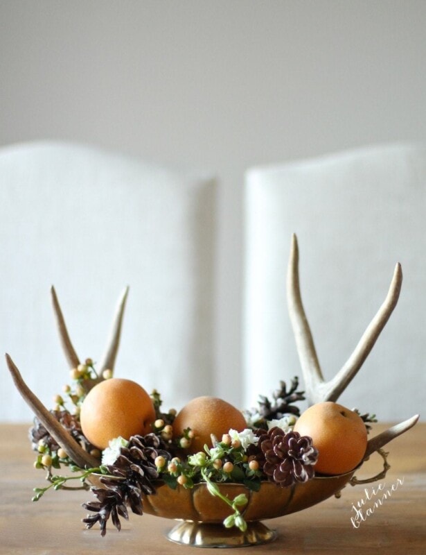 A DIY Thanksgiving centerpiece of a brass bowl full of fruit, pine cones and antlers