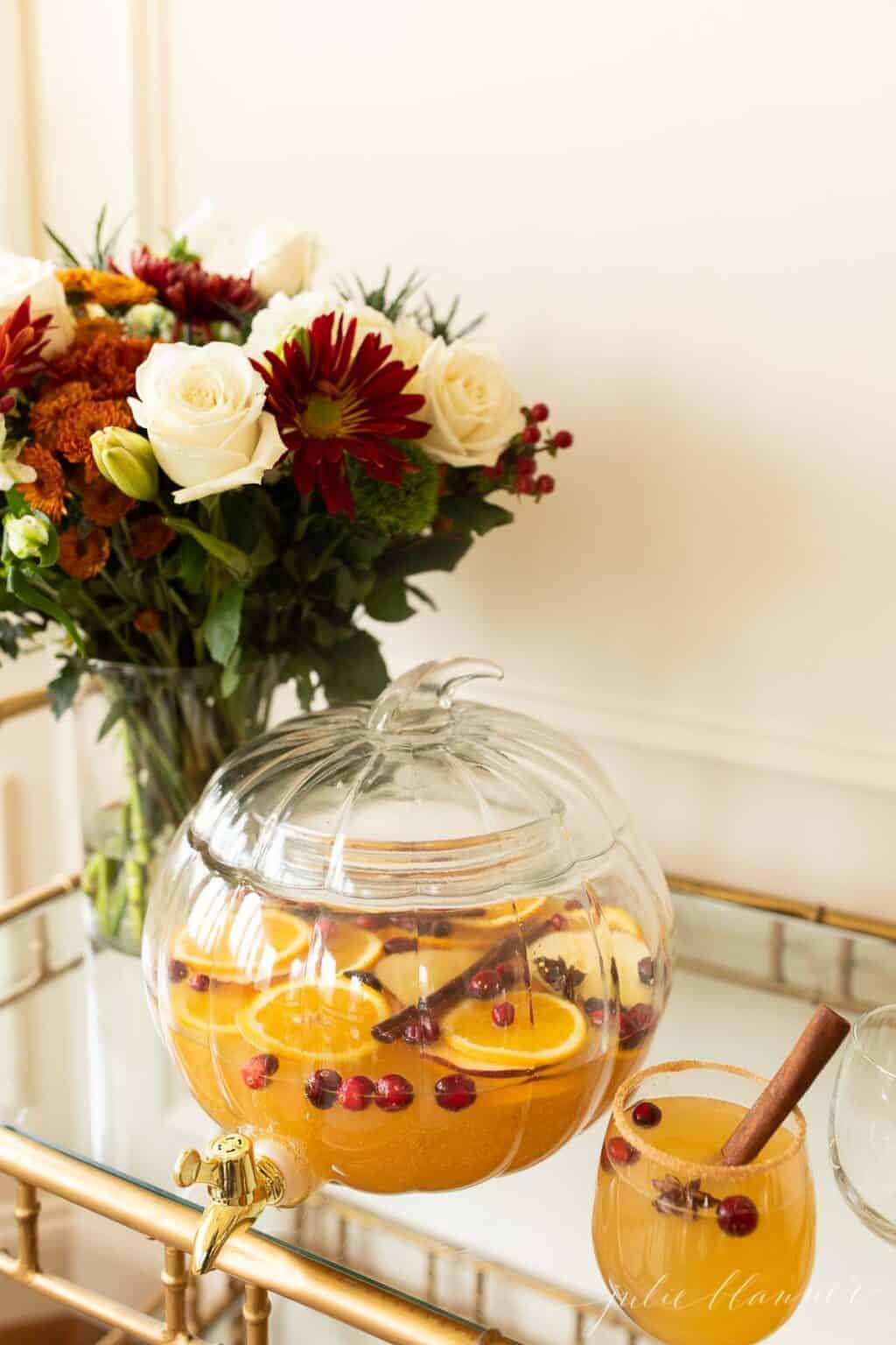 A clear glass pumpkin drink dispenser filled with apple cider sangria, flowers in the background.