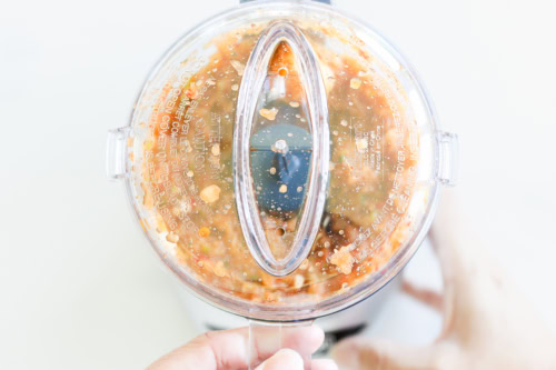 Top view of a blender filled with blended ingredients for chipotle vinaigrette, including bits of food visible through the clear lid, against a light background.