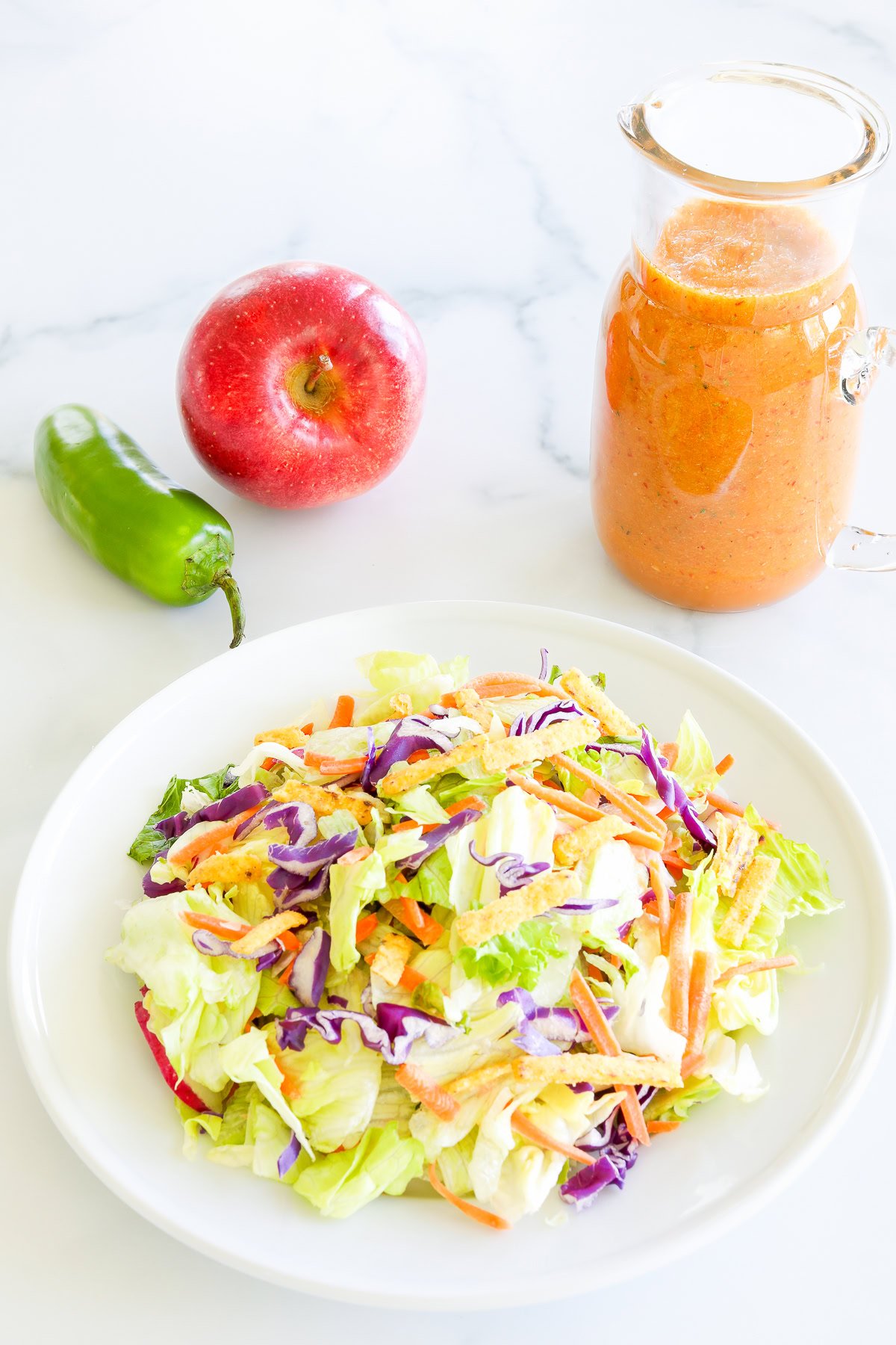 A vibrant salad with mixed vegetables on a white plate, a red apple, and a jar of chipotle salad dressing beside a green jalapeño on a marble surface.