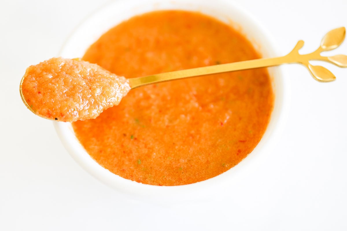 A golden spoon lifting tomato soup from a white bowl, with a visible mix of herbs and chipotle vinaigrette.