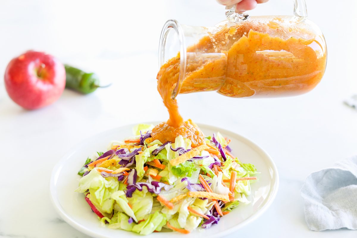 Pouring chipotle vinaigrette from a glass jar onto a colorful mixed salad on a white plate, with an apple and bell pepper visible in the background.