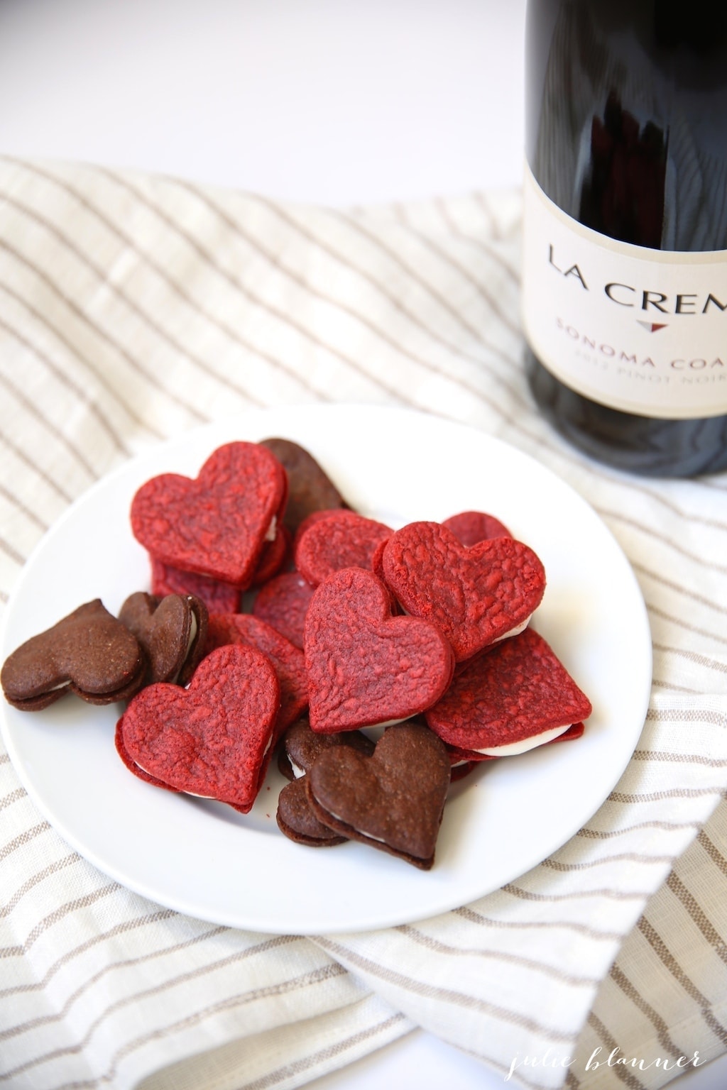 heart shaped red velvet sandwich cookies on a plate with chocolate oreos next to a bottle of wine