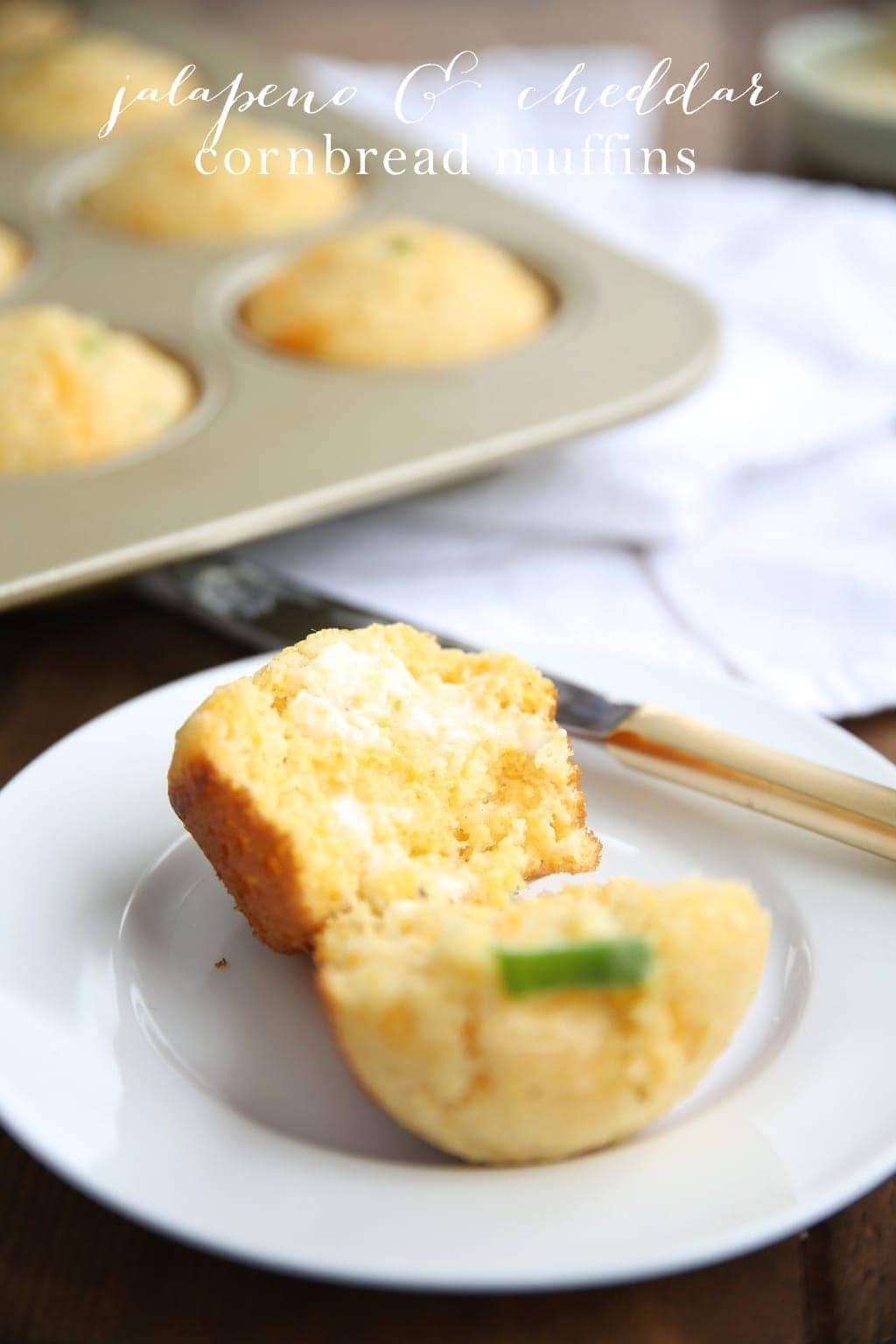 A cornbread muffin on a white plate, cut in half and buttered, with a tray of cornbread muffins in the background