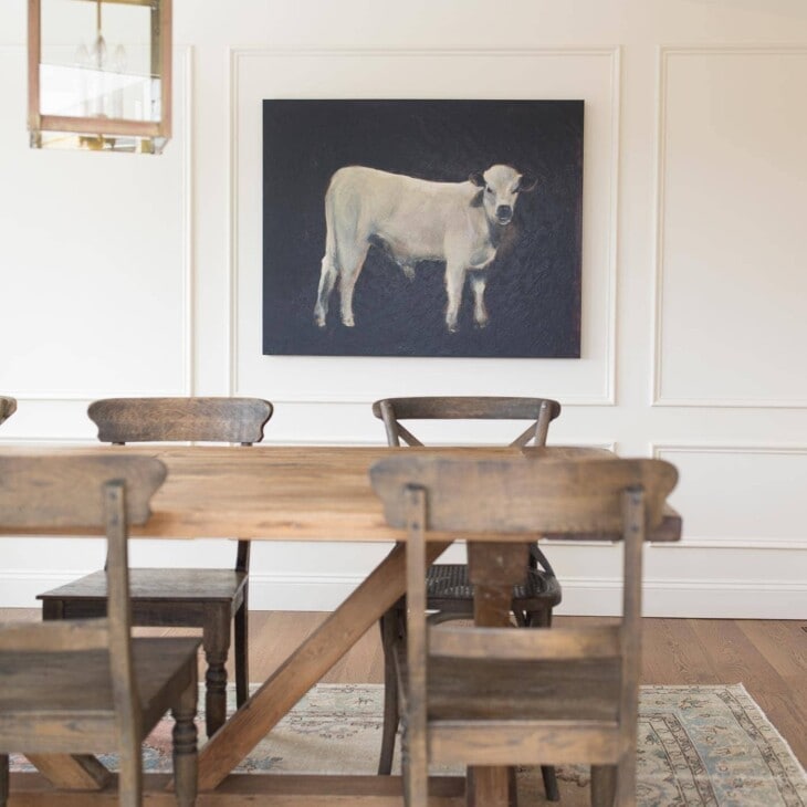 farmhouse table on vintage rug with cream colored walls large cow painting and brass lantern pendant