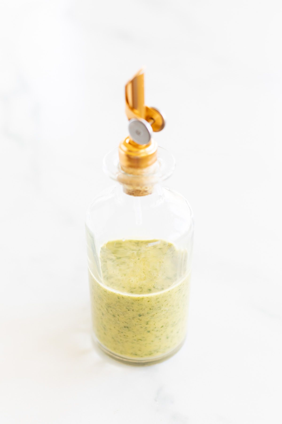 A glass bottle with a gold spout, full of cilantro lime dressing. Placed on a white marble countertop.