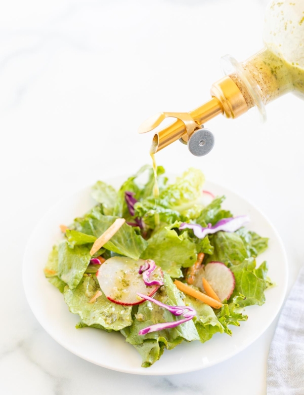 A glass bottle of cilantro lime dressing being poured over a green salad on a white plate.