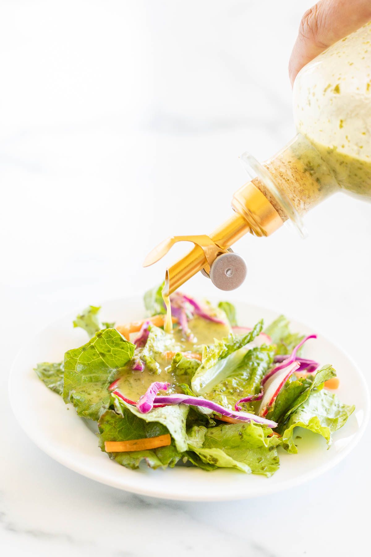 A glass bottle of cilantro lime dressing being poured over a green salad on a white plate.