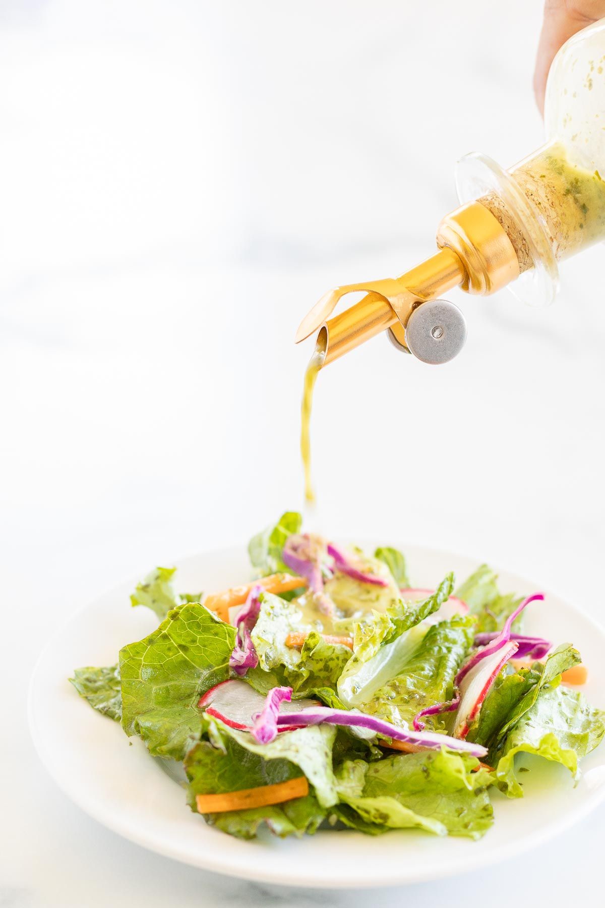 A glass bottle of cilantro lime dressing being poured over a green salad on a white plate.