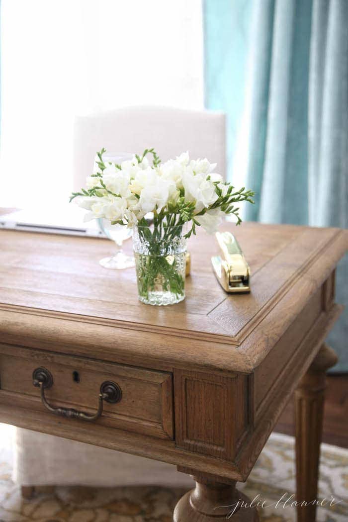 A wooden desk in a home office, vase of flowers and gold stapler on top.