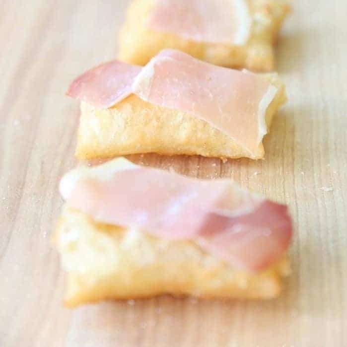 A row of gnocco fritto on a wooden surface, topped with prosciutto