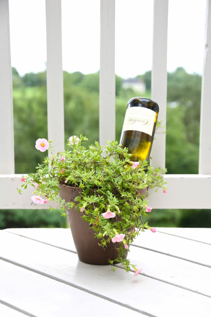 A vase of flowers on a white porch with a wine bottle inside. 