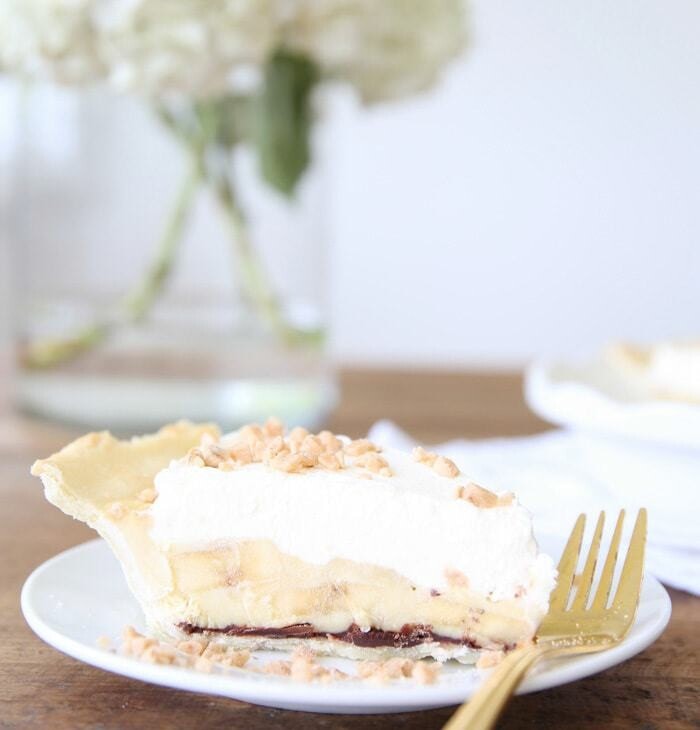 slice of easy banana cream pie recipe on white plate with gold fork with white flowers in glass vase in background