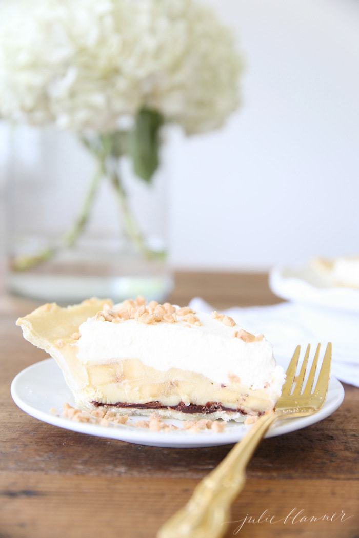 slice of easy banana cream pie recipe on white plate with gold fork with white flowers in glass vase in background