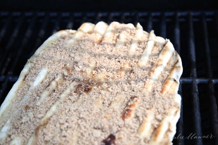 Pizza dough spread out on an outdoor grill, topped with cinnamon and brown sugar