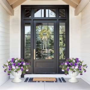 A black-framed glass front door with a wreath, flanked by two white porch planters filled with purple flowers, and a doormat on a striped rug.
