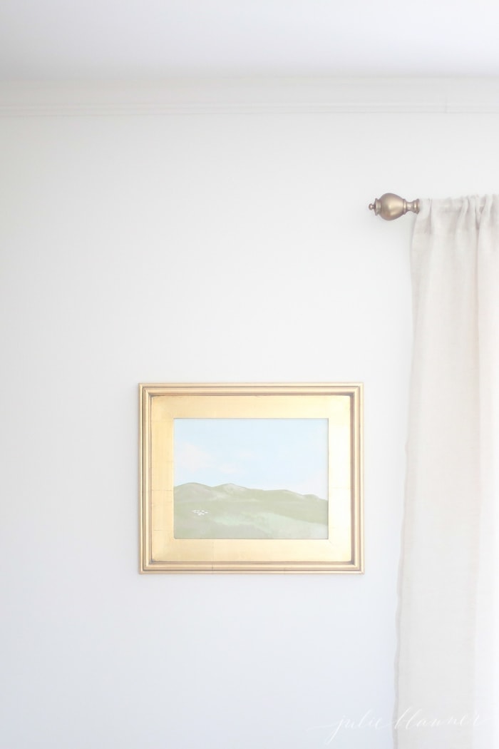 A white wall with a framed oil painting in a gold frame.