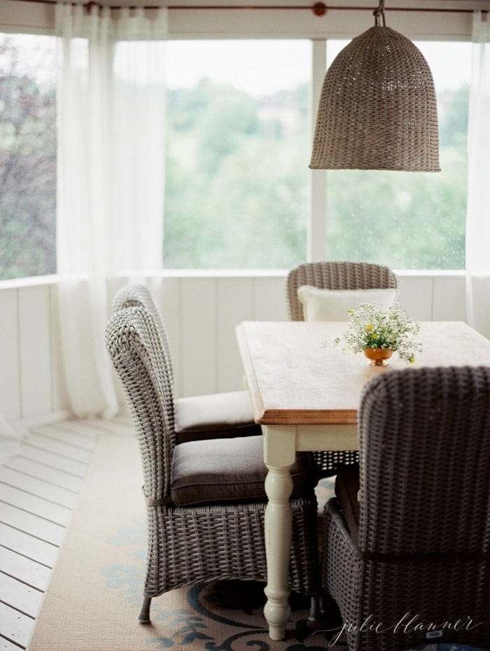 A table with woven chairs and light fixture above it in a screened in porch. 