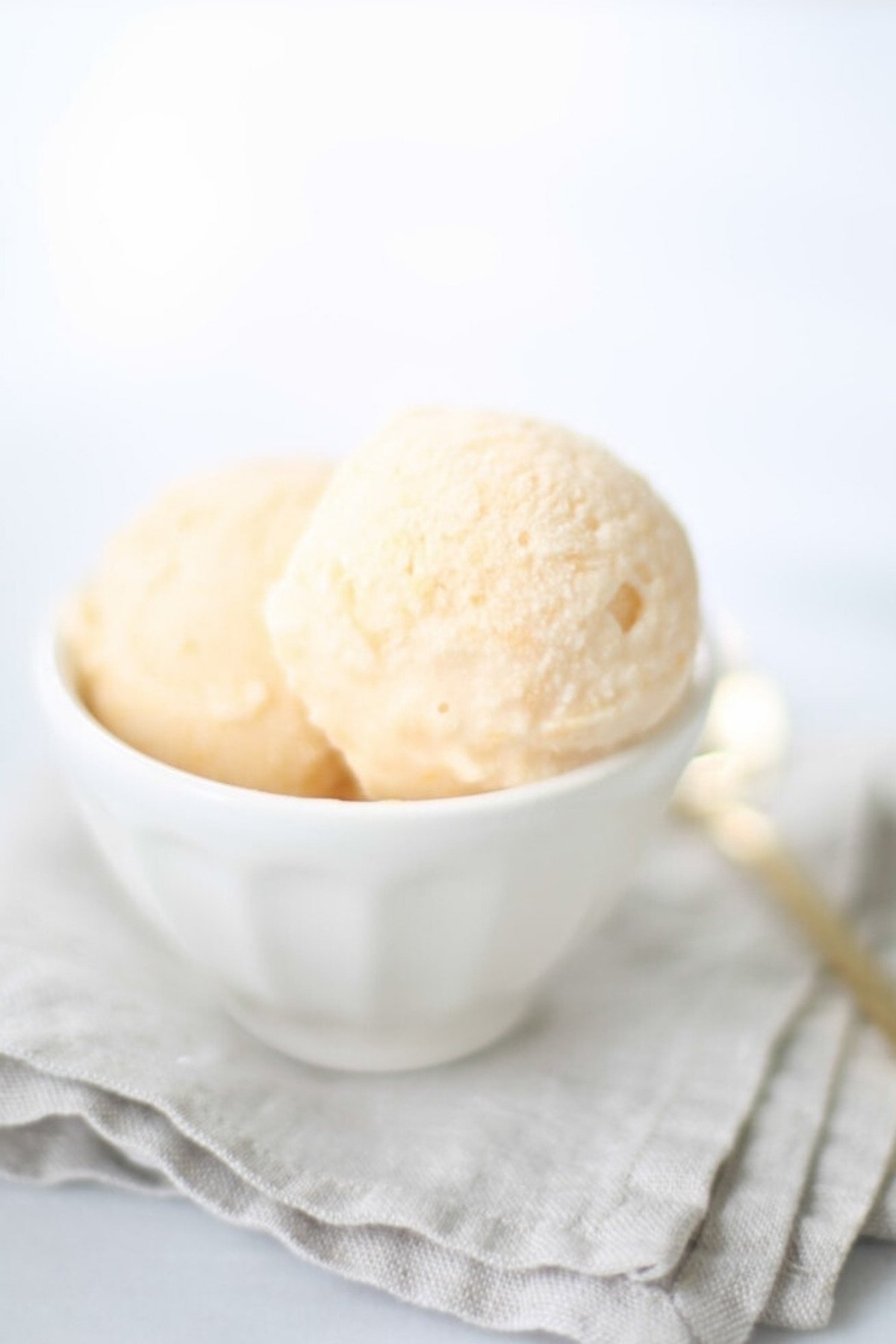 A white bowl contains two scoops of no churn peach ice cream, placed on a folded beige napkin with a gold spoon beside it.