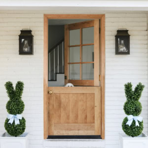 A wooden Dutch door is half open in a white brick house. Two topiary bunnies with blue bows sit in porch planters on either side of the entrance, while a dog peeks over the lower door.