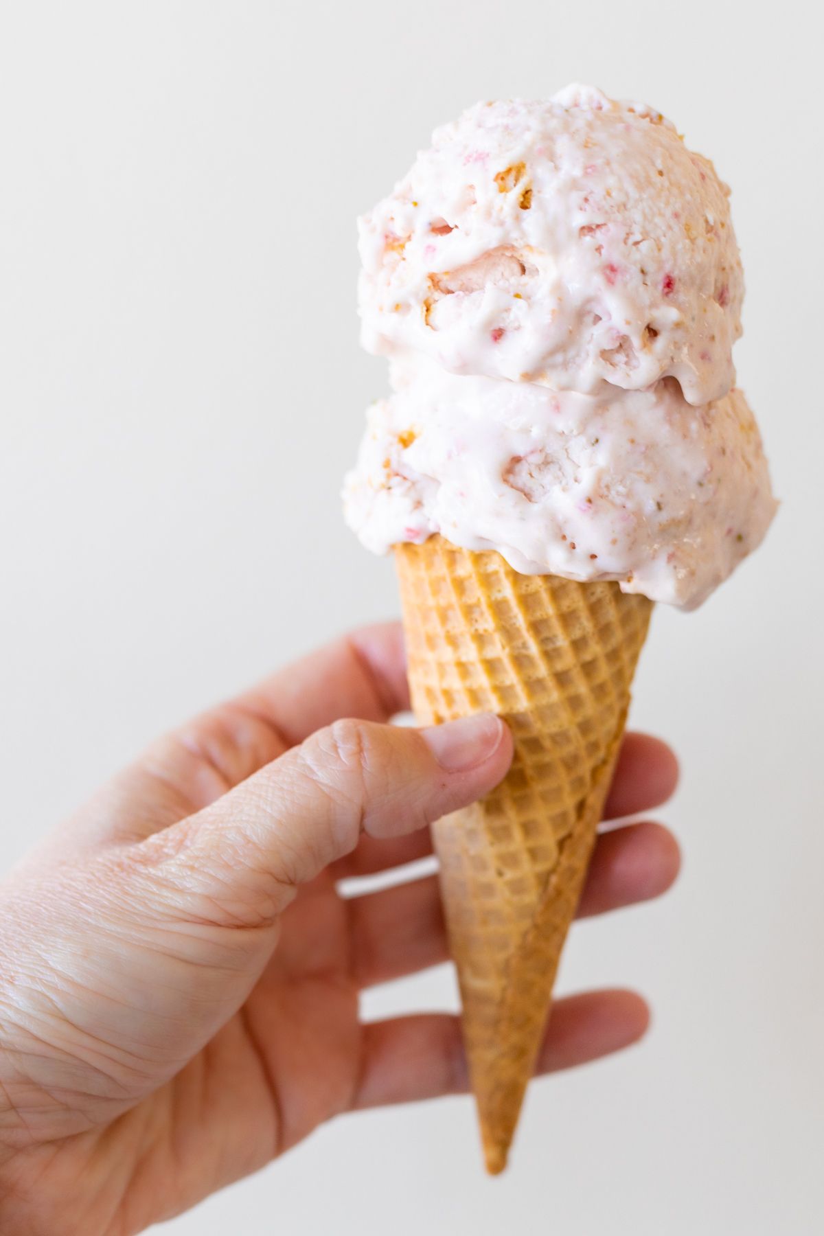 A hand holding a cone of strawberry cheesecake ice cream.