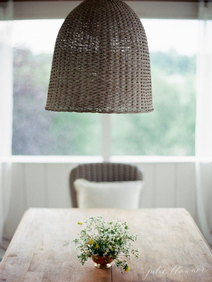 A table with woven chairs and light fixture above it in a screened in porch. 