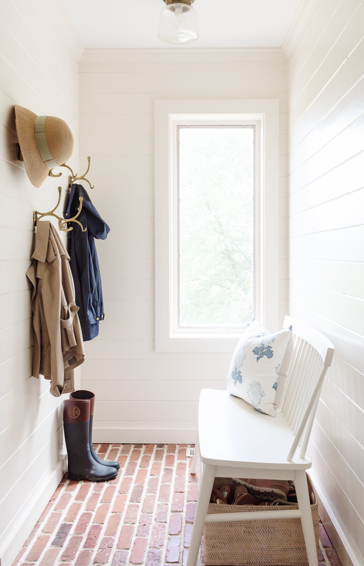 A white mudroom with brick floors, a white bench and brass hooks on the wall.
