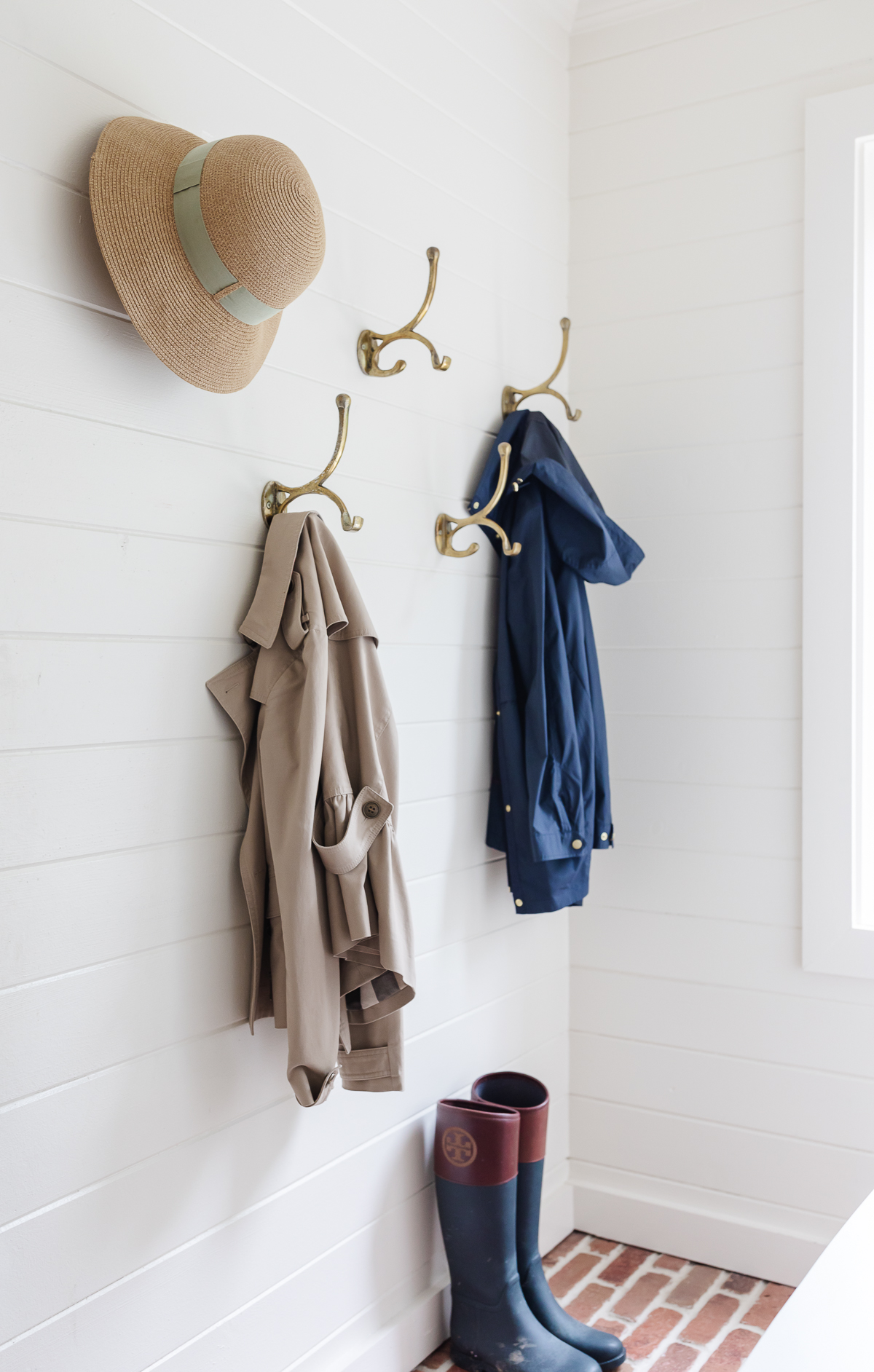 A white mudroom with brick floors and brass hooks on the wall.