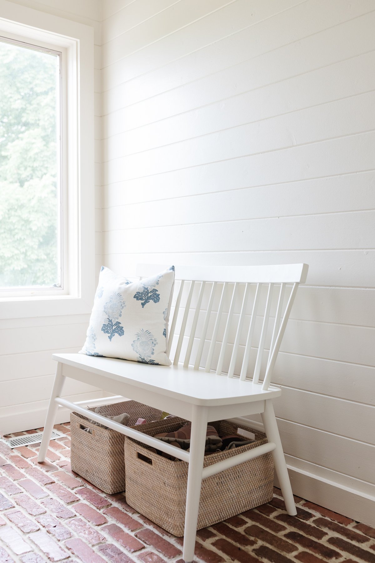 A white mudroom with brick floors, a white bench and brass hooks on the wall.