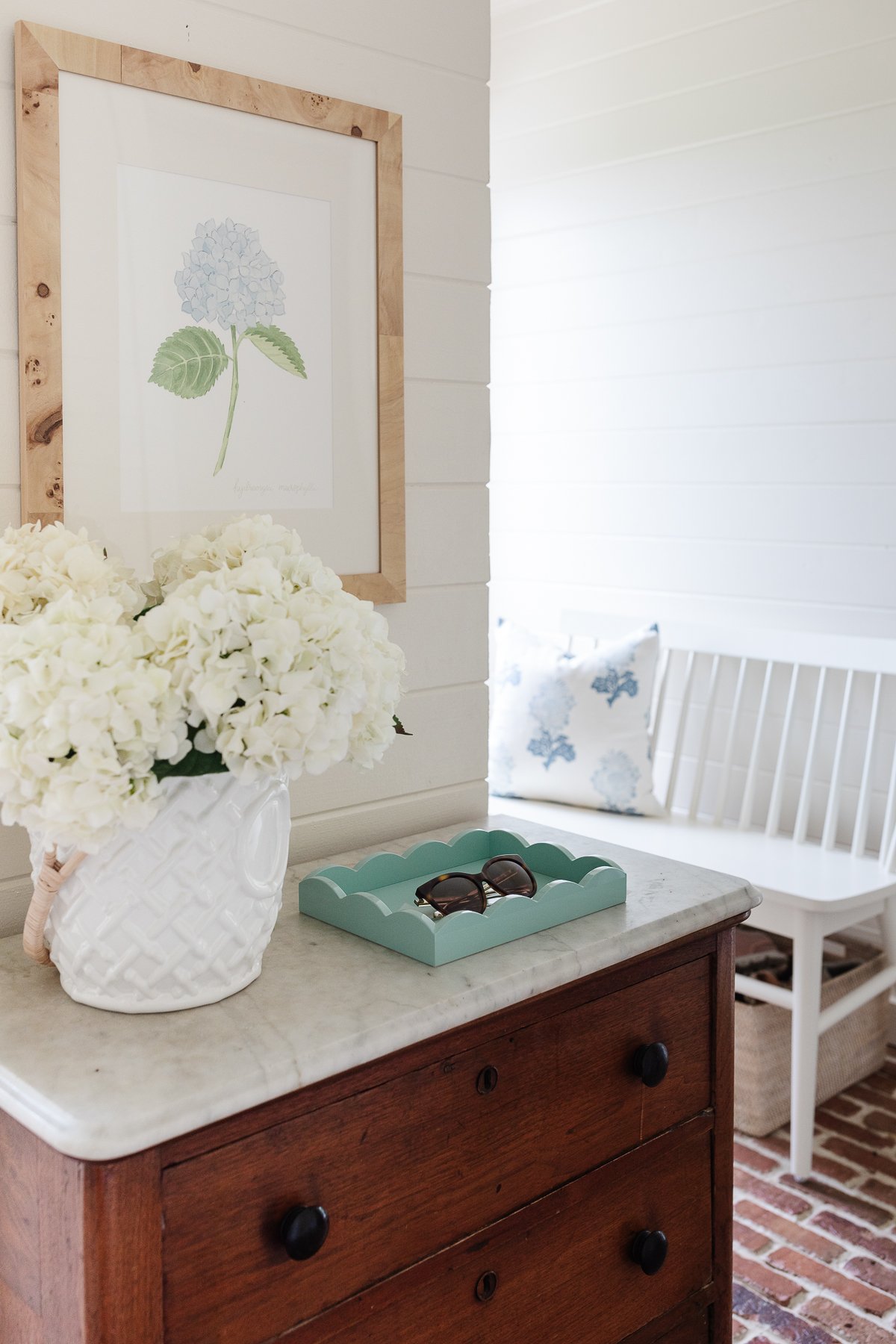 A white mudroom with brick floors, a white bench and brass hooks on the wall.