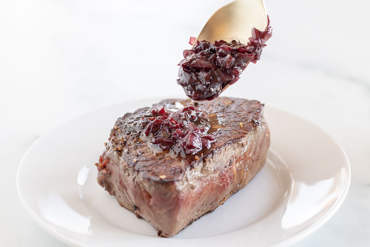 A steak on a white plate, with a gold spoon full of red wine jus being drizzled over it