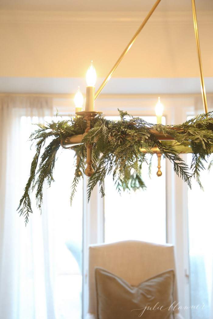 A brass chandelier dressed up with winter decor christmas greens over a dining table.
