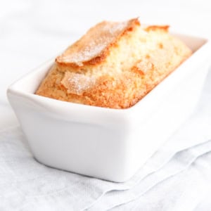 A loaf of freshly baked sweet bread in a white ceramic baking dish, placed on a light blue cloth.
