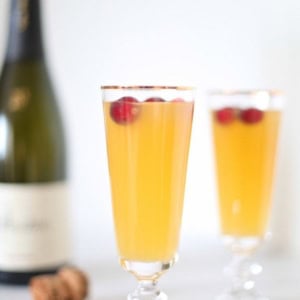Two champagne flutes filled with a yellow-orange pear prosecco cocktail garnished with cranberries, with a bottle of champagne and cork in the background.