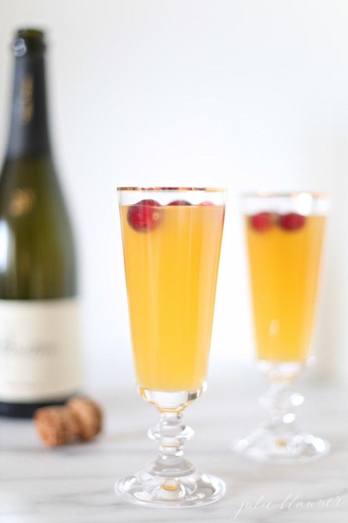 Two champagne flutes filled with a yellow-orange pear prosecco cocktail garnished with cranberries, with a bottle of champagne and cork in the background.