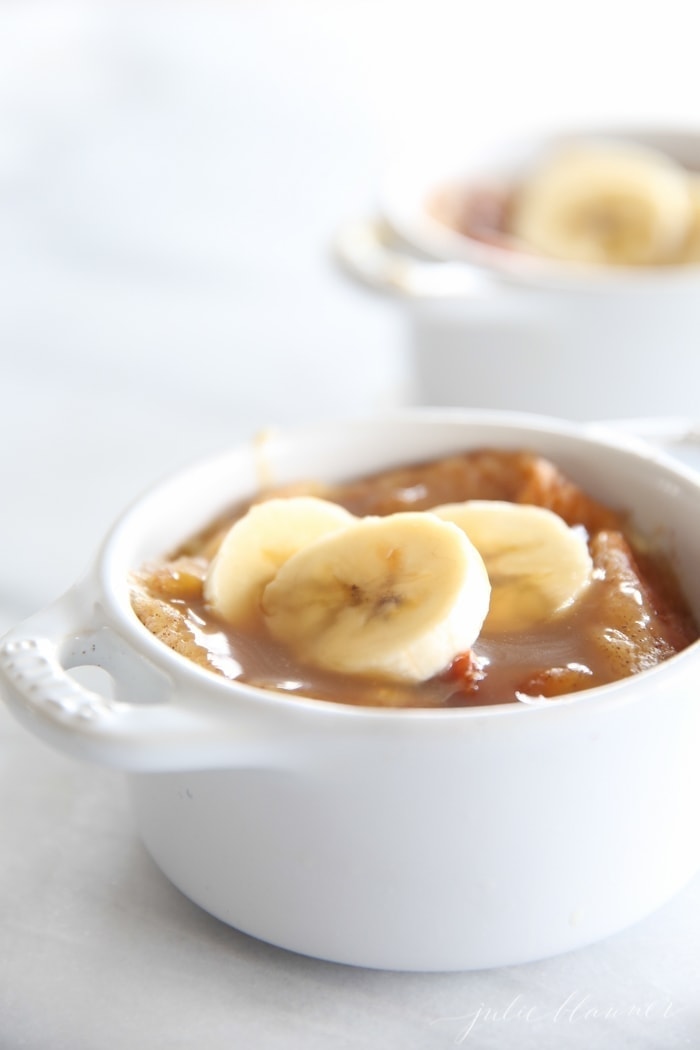 easy bread pudding topped with bourbon sauce and banana slices in two white individual baking dishes