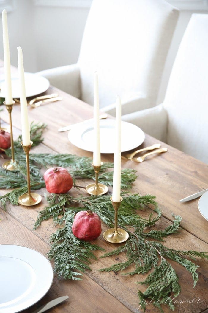 A Christmas candle centerpiece set on a wood table, featuring brass taper holders and white taper candles, pomegranates and cedar. 