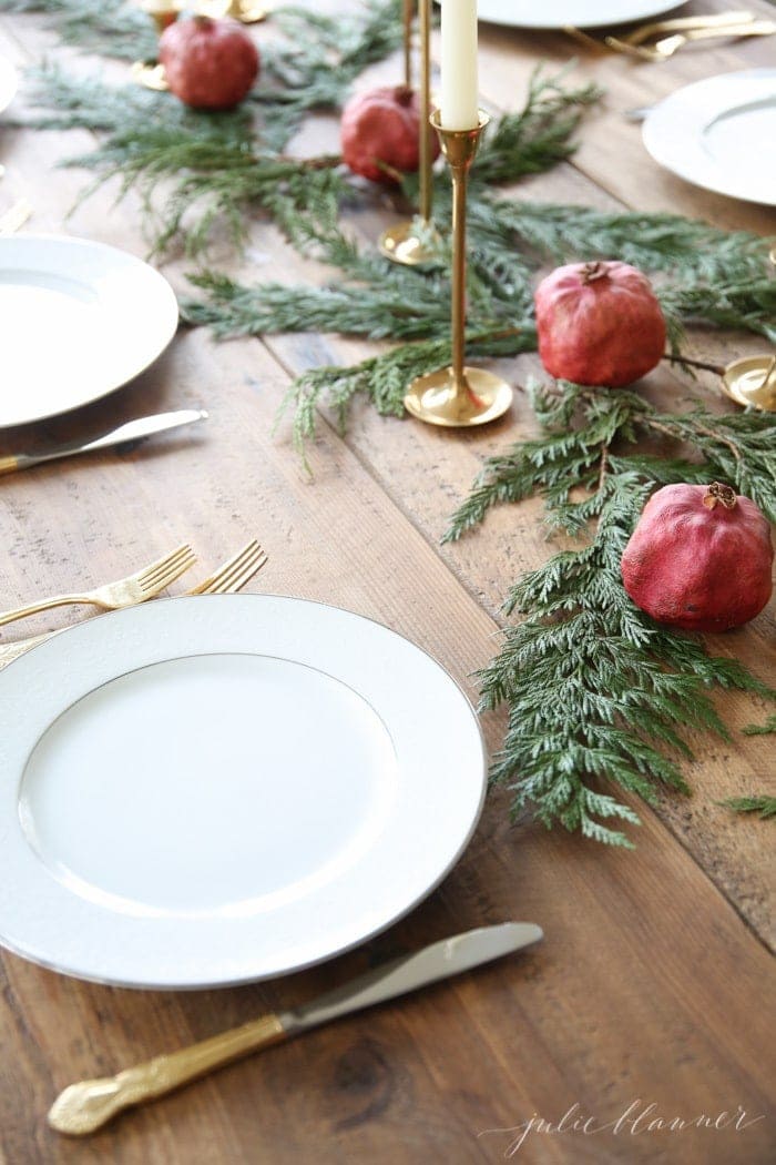 A Christmas candle centerpiece set on a wood table, featuring brass taper holders and white taper candles, pomegranates and cedar. 