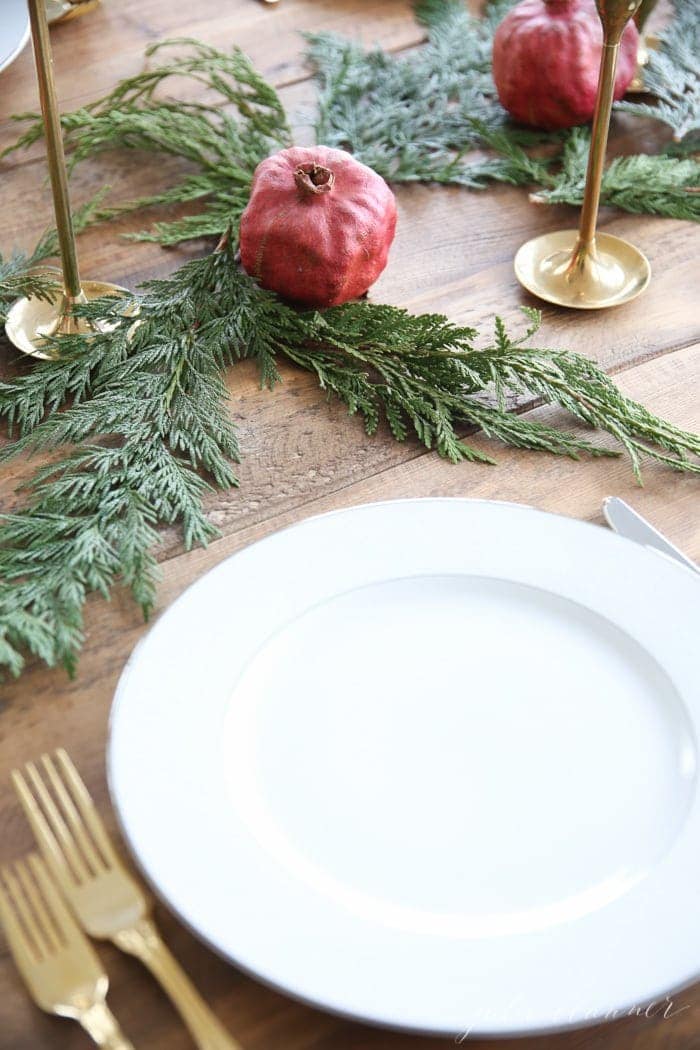 A Christmas candle centerpiece set on a wood table, featuring brass taper holders and white taper candles, pomegranates and cedar. 