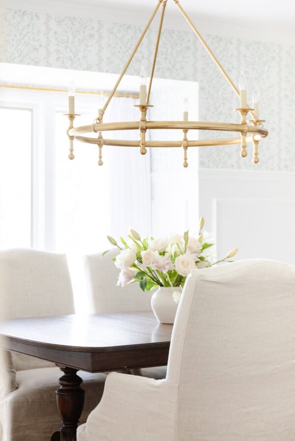 A dark wood dining table with a white floral arrangement is surrounded by upholstered chairs under a gold chandelier in a bright room—ideal for those seeking inspiration on how to clean linen chair upholstery beautifully.