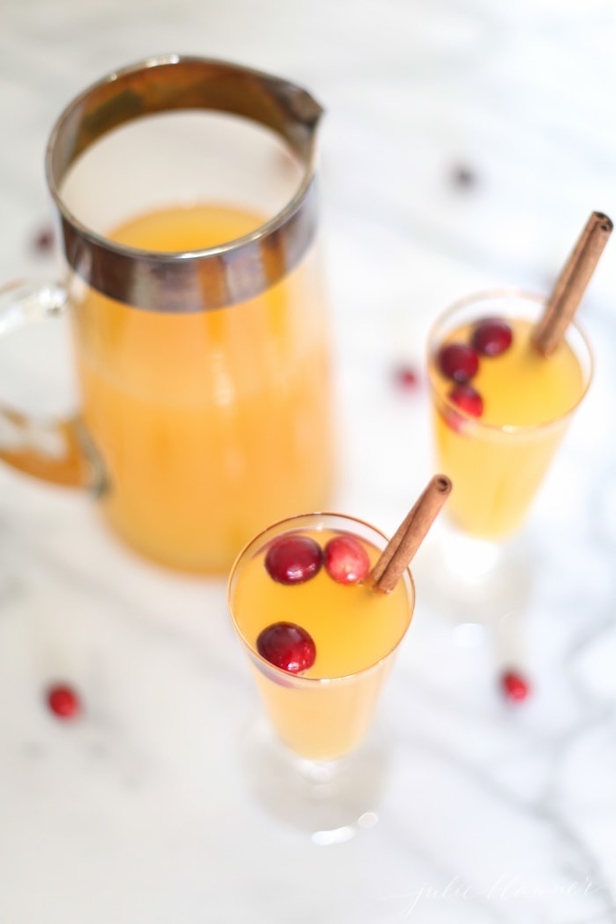 A pitcher and two glasses of yellow-orange pear prosecco cocktail, garnished with cranberries and cinnamon sticks, are placed on a white marble surface.