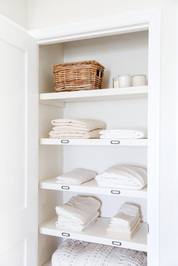 A linen closet with neatly folded white sheets, a woven basket on the top shelf, and white candles creates a serene and organized linen closet space.