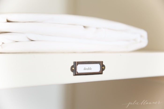 A hall linen closet organization project with labels on each shelf and white sheets.