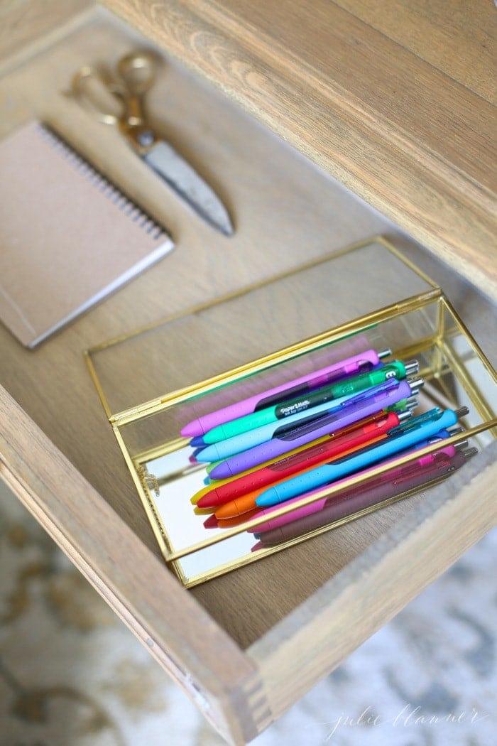 Looking inside a wood desk that is carefully organized, pens in a pretty glass box.