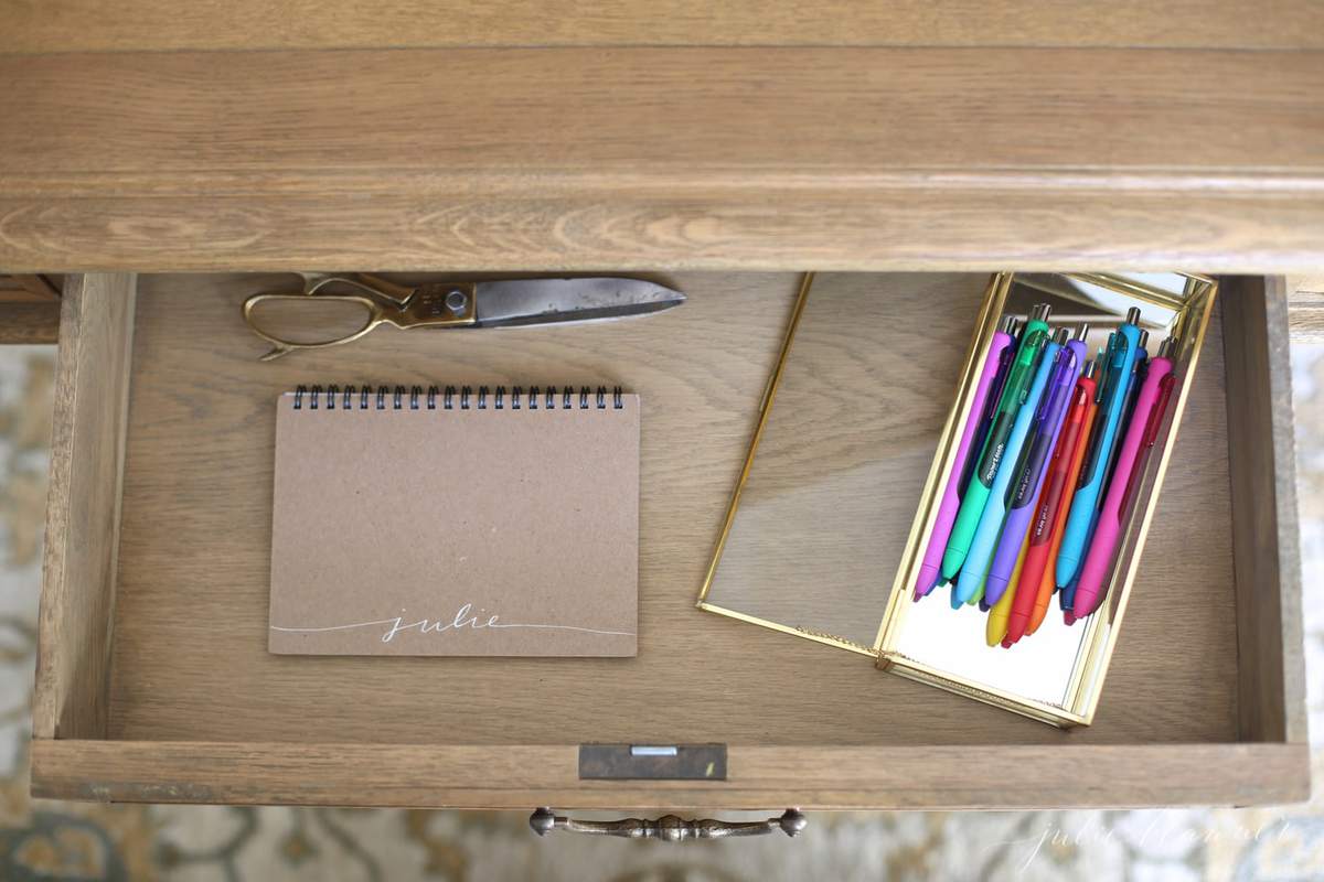 Inside a clean wooden desk drawer, organized notebook, scissors and a gold case full of pens.