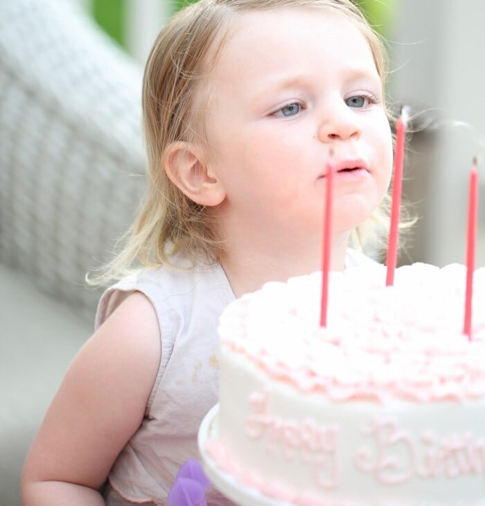 A little girl with blonde hair, blowing out tall pink candles on a white birthday cake