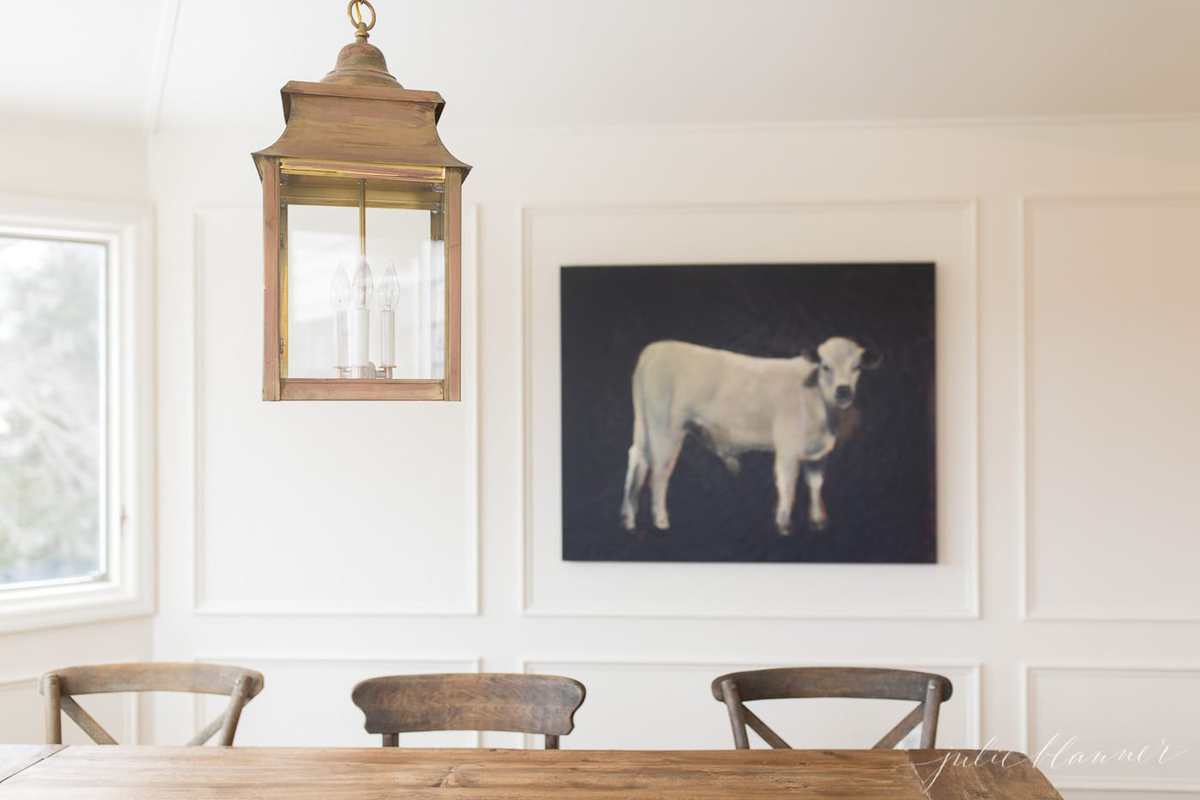 A white breakfast nook room off a kitchen, with a farm table, brass lantern and a cow painting in the background.