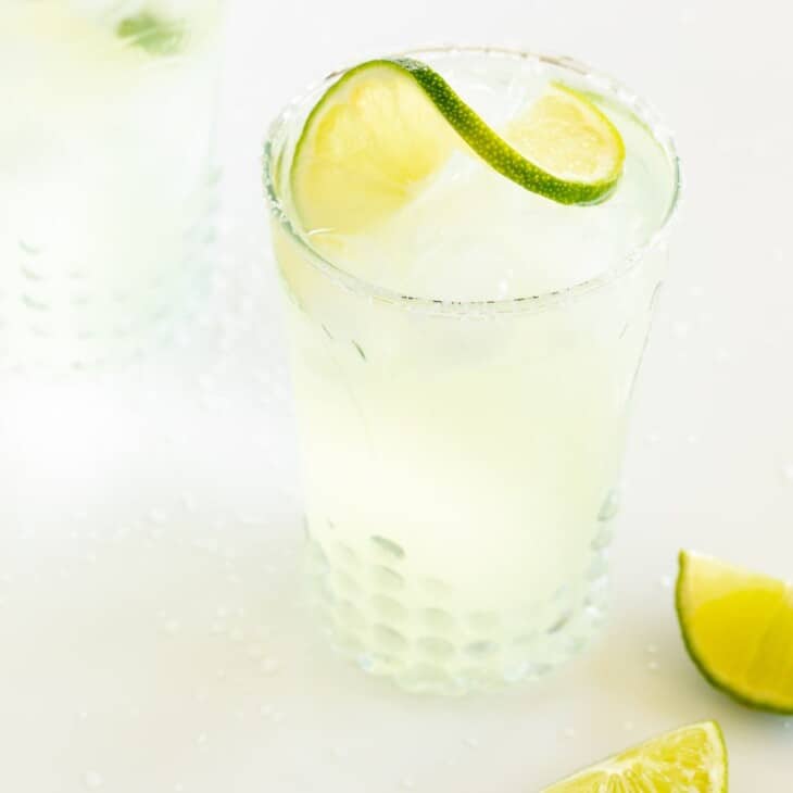 A clear glass with a homemade margarita, rimmed with salt and a slice of lime on a white background, lime wedges to the side.
