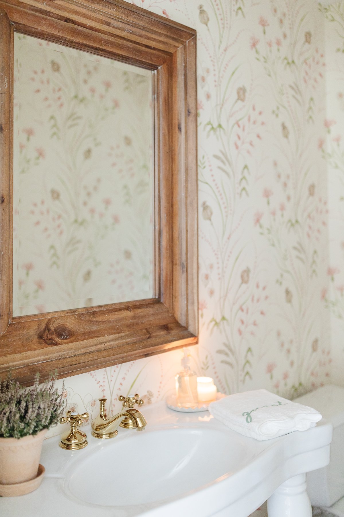 A wallpapered powder room with a wood mirror and white pedestal sink.