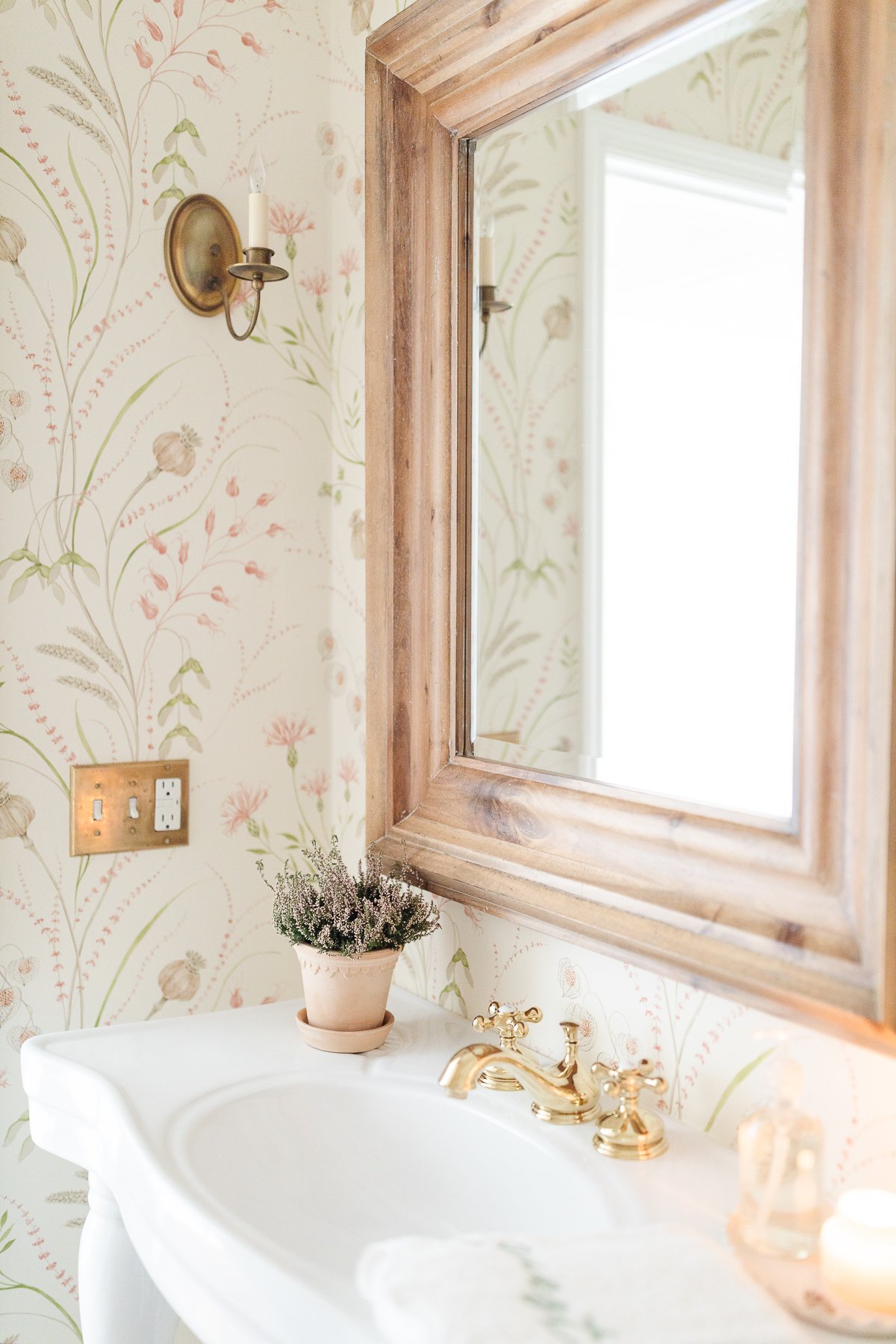 A wallpapered powder room with a wood mirror and white pedestal sink.